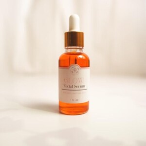 K&ouml;nnte beinhalten: Eine durchsichtige Glasflasche mit goldenem Deckel und Tropfer, gef&uuml;llt mit einer hellorangen Fl&uuml;ssigkeit. Die Flasche hat ein wei&szlig;es Etikett mit dem Text "GLOW Facial Serum" und "1 FL OZ".