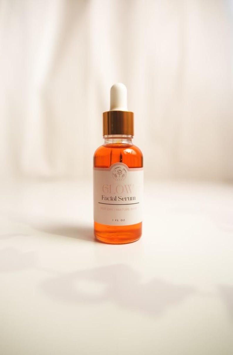 K&ouml;nnte beinhalten: Eine durchsichtige Glasflasche mit goldenem Deckel und Tropfer, gef&uuml;llt mit einer hellorangen Fl&uuml;ssigkeit. Die Flasche hat ein wei&szlig;es Etikett mit dem Text "GLOW Facial Serum" und "1 FL OZ".