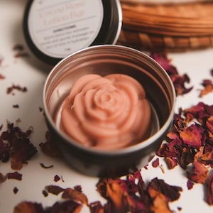 May include: A rose-shaped pink soap bar in a silver tin container. The soap is surrounded by dried rose petals.