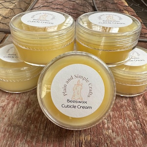 Op de afbeelding: Een verzameling van vijf kleine, doorzichtige plastic potjes gevuld met gele nagelriemcrème. Elk potje heeft een wit etiket met de tekst "Plain and Simple Crafts" en "Beeswax Cuticle Cream". De potjes staan op een houten oppervlak.