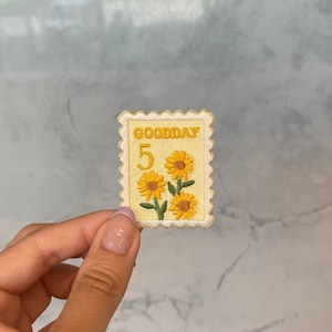 May include: A yellow and white embroidered patch with the word "GOODDAY" and the number "5" in the centre. The patch features three yellow sunflowers with green stems and leaves.