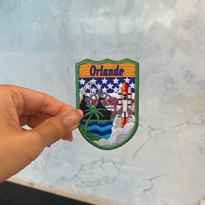 May include: Embroidered patch with "Orlando" text. The design includes a rocket launch, Ferris wheel, palm trees, and a starry background. The patch has a green border. The design is in shades of blue, orange, and green.