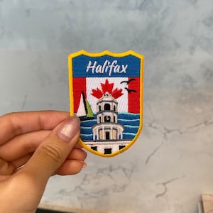 Puede incluir: Un parche bordado en amarillo, azul y rojo con la palabra "Halifax" en letras blancas. El parche presenta un velero blanco, una bandera canadiense roja con una hoja de arce y un edificio blanco con una torre de reloj. El edificio está rodeado de olas azules.