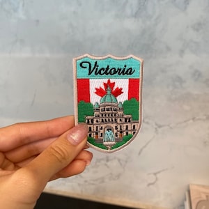Könnte beinhalten: Ein gestickter Aufnäher in Weiß, Grün und Rot mit dem Text "Victoria" und einer Darstellung der Parlamentsgebäude in Victoria, British Columbia, Kanada.
