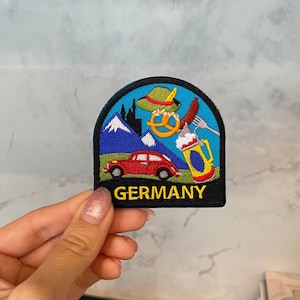 Peut inclure: Écusson brodé avec une bordure bleue, représentant une voiture rouge, des montagnes, un bretzel, une chope de bière et un chapeau traditionnel. Le mot "GERMANY" est en jaune.