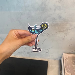 Peut inclure: Un patch brodé représentant un verre à martini avec un quartier de citron. Le verre est rempli de liquide bleu et a une paille qui en sort.