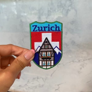 以下が含まれることがあります： 「チューリッヒ」という言葉とスイスの国旗が刺繍された青、緑、赤、白の刺繍パッチ。パッチには、赤い屋根と白い窓のある小さな家も描かれています。