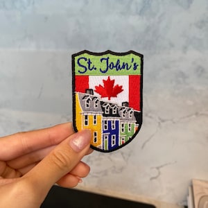 May include: An embroidered patch featuring the text "St. John's" in green, yellow, red and blue. The patch also includes a Canadian flag with a red maple leaf and a row of colourful houses.