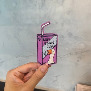 Op de afbeelding: Een geborduurde patch van een roze en witte sapdoos met een rietje en een gans op de voorkant. De sapdoos zegt "Silly Goose Juice".
