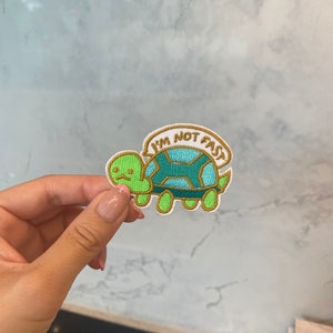 May include: Embroidered patch depicting a cartoon turtle in green and teal, with the text "I'M NOT FAST" in a banner. The patch has a white border and is designed for application on clothing or accessories.