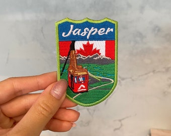 Puede incluir: Parche bordado en verde, azul y rojo con la palabra "Jasper" en letras blancas. El parche presenta un teleférico rojo con una bandera canadiense en la parte superior, que sube una montaña verde con un río azul.