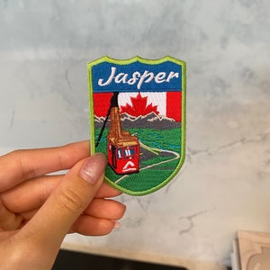 May include: Green, blue, and red embroidered patch with the word "Jasper" in white lettering. The patch features a red cable car with a Canadian flag on top, traveling up a green mountain with a blue river.
