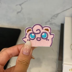 May include: Pink and purple embroidered patch of a cartoon character with blue eyes and a pink nose. The character has a fluffy, cloud-like appearance.