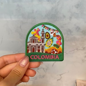 Puede incluir: Un parche bordado verde, azul y rosa con una iglesia, un balón de fútbol y la palabra "COLOMBIA" en letras rosas.