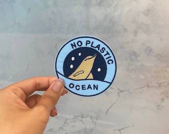 Può includere: Un cerotto rotondo blu e nero con il testo "NO PLASTIC OCEAN" e un'immagine di un uccello dei cartoni animati.