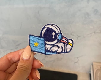 May include: A blue and white embroidered patch of an astronaut sitting in front of a laptop with a yellow star on the screen.