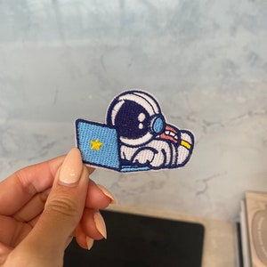 Peut inclure: Un patch brodé bleu et blanc représentant un astronaute assis devant un ordinateur portable avec une étoile jaune sur l'écran.