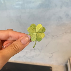 May include: A four-leaf clover patch in shades of green, with detailed embroidery. The clover has four distinct heart-shaped leaves and a small stem. The patch is likely for clothing or accessories.