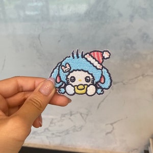 May include: An embroidered patch featuring a cartoon character in a blue and white design, wearing a red and white Santa hat. The character has a yellow dummy in its mouth.