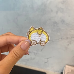 Könnte beinhalten: Ein gestickter Aufnäher mit dem Hinterteil eines Cartoon-Hundes. Der Hund ist gelb und braun, trägt ein weißes Hemd und hat einen braunen Schwanz.
