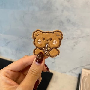 May include: Brown embroidered patch of a cartoon bear wearing a brown hat and drinking from a straw. The bear has pink cheeks and a brown nose.