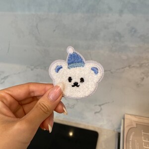 May include: White fuzzy bear applique with a blue knit hat. The bear has black eyes and a black nose.