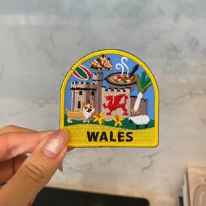 Può includere: Una toppa ricamata gialla e blu con un drago gallese, un castello, un porro, una bandiera gallese, un corgi, una pecora e una ciotola di stufato gallese. La toppa dice "WALES" in lettere nere.