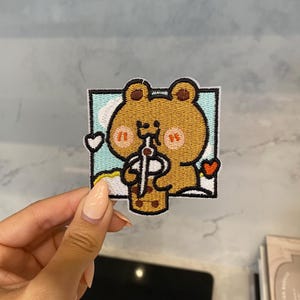 Könnte beinhalten: Gestickter Aufnäher mit einem braunen Bären-Cartoon-Charakter, der eine braune und weiße Boba-Teetasse hält. Der Bär trägt ein braunes Hemd und hat eine rosa Nase und Wangen. Der Aufnäher ist von einem blauen und weißen Rand mit Wolken und Herzen umgeben.