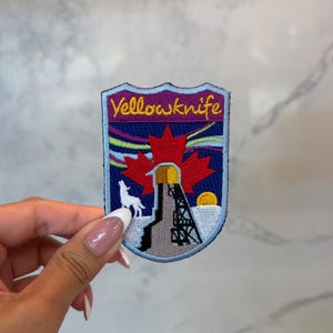 Peut inclure: Écusson brodé avec le mot "Yellowknife" en jaune. Le motif comprend une feuille d'érable rouge, un loup blanc et une structure avec un soleil jaune. Le fond présente des couleurs violettes, bleues et jaunes.