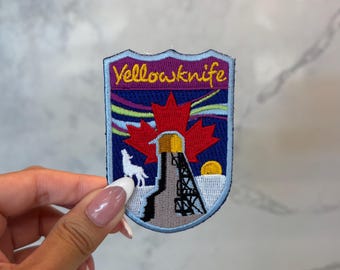 May include: Embroidered patch with the word "Yellowknife" in yellow. The design includes a red maple leaf, a white wolf, and a structure with a yellow sun. The background features purple, blue, and yellow colours.