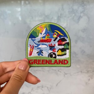 May include: A green, blue, red, and yellow embroidered patch with a scene of a snowy landscape, houses, and a penguin. The text "GREENLAND" is written in red on the bottom of the patch.