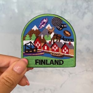 May include: An embroidered patch featuring a winter scene of houses, mountains, and a reindeer. The patch is green, blue, and red and says "FINLAND" in black letters.
