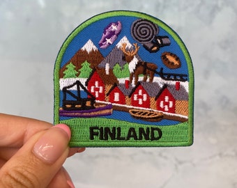 Può includere: Una toppa ricamata verde, blu e rossa con una scena invernale di case, montagne e una renna. La toppa dice "FINLANDIA" in lettere nere.