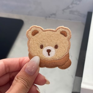 Peut inclure: Un patch brodé en forme d'ours brun et beige avec un nez blanc et des yeux bruns. L'ours porte une écharpe brune.