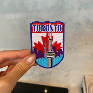 Può includere: Una toppa ricamata blu e bianca con la parola "Toronto" e una rappresentazione della CN Tower e una bandiera canadese.
