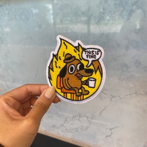 May include: An embroidered patch featuring a cartoon dog sitting in a chair with a cup of coffee. The dog is wearing a hat and has a speech bubble that says "This is fine".