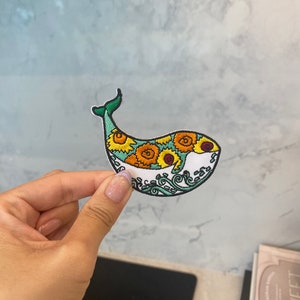 Peut inclure: Un écusson brodé de baleine blanche avec une queue verte et un motif de tournesols jaunes sur son corps. La baleine est entourée d'un motif de vagues bleues et vertes.