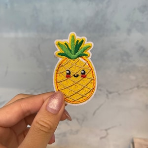 Op de afbeelding: Een geborduurde ananas-patch met een geel lichaam, een groene bovenkant en een glimlachend gezicht. De ananas bevindt zich op een witte achtergrond.
