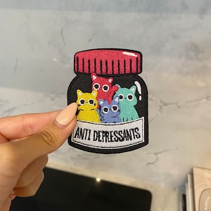 Pode incluir: Patch bordado com um frasco de comprimidos preto com tampa vermelha. Dentro do frasco estão cinco gatos de desenho animado coloridos. O patch diz "ANTI DEPRESSANTS" em texto branco.