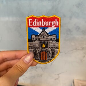 May include: Embroidered patch with the word "Edinburgh" above a castle illustration. The shield-shaped patch has a red and blue border, and a yellow outline. The castle is depicted in gray with a brown door, a souvenir item.