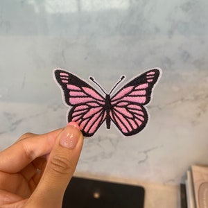 May include: Embroidered butterfly patch featuring pink wings and black detailing. The patch has a white border and intricate wing patterns. This decorative patch can be ironed or sewn onto fabric items like clothing or bags.