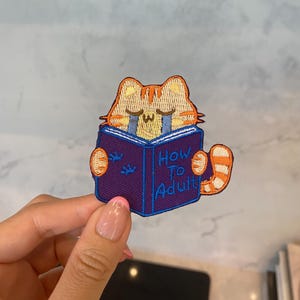 Puede incluir: Un parche bordado para planchar con un gato de dibujos animados leyendo un libro titulado "How to Adult". El gato es naranja y blanco con una expresión triste y lleva una camisa azul. El libro es morado con texto azul y blanco.