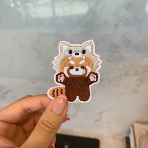 Peut inclure: Un patch brodé représentant un panda roux debout sur ses pattes arrière, les pattes levées en l'air. Le patch est brun et blanc avec un contour blanc et mesure environ 5 cm de haut.