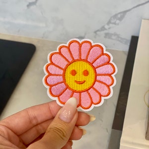 Puede incluir: Un parche bordado de una flor rosa y naranja con una carita sonriente amarilla en el centro.