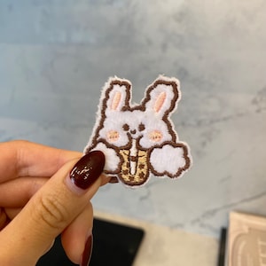Puede incluir: Conejo blanco bordado sosteniendo una taza de té boba marrón y blanca. El conejo tiene orejas rosas y un contorno marrón.