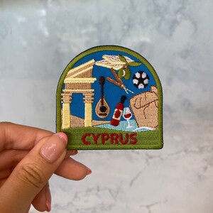 May include: Embroidered patch featuring a Cyprus theme. The patch has a blue background with images of a column, a musical instrument, a bottle of wine, and a soccer ball. The word "CYPRUS" is embroidered in red.