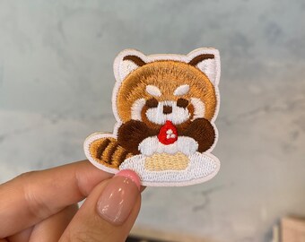 Puede incluir: Un parche bordado con un panda rojo con pelaje marrón, vientre blanco y una fresa roja en sus patas. El parche está sobre un fondo blanco.