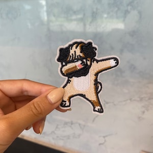 May include: An embroidered iron-on patch of a pug dog doing the dab dance, in brown and black.