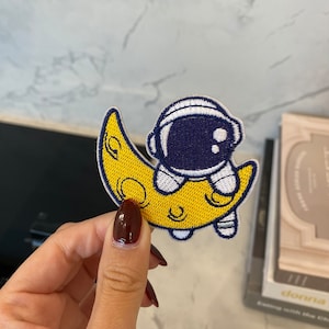 May include: A yellow crescent moon with a white and blue astronaut sitting on top. The astronaut has a white helmet and a blue spacesuit.