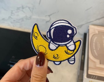 May include: A yellow crescent moon with a white and blue astronaut sitting on top. The astronaut has a white helmet and a blue spacesuit.